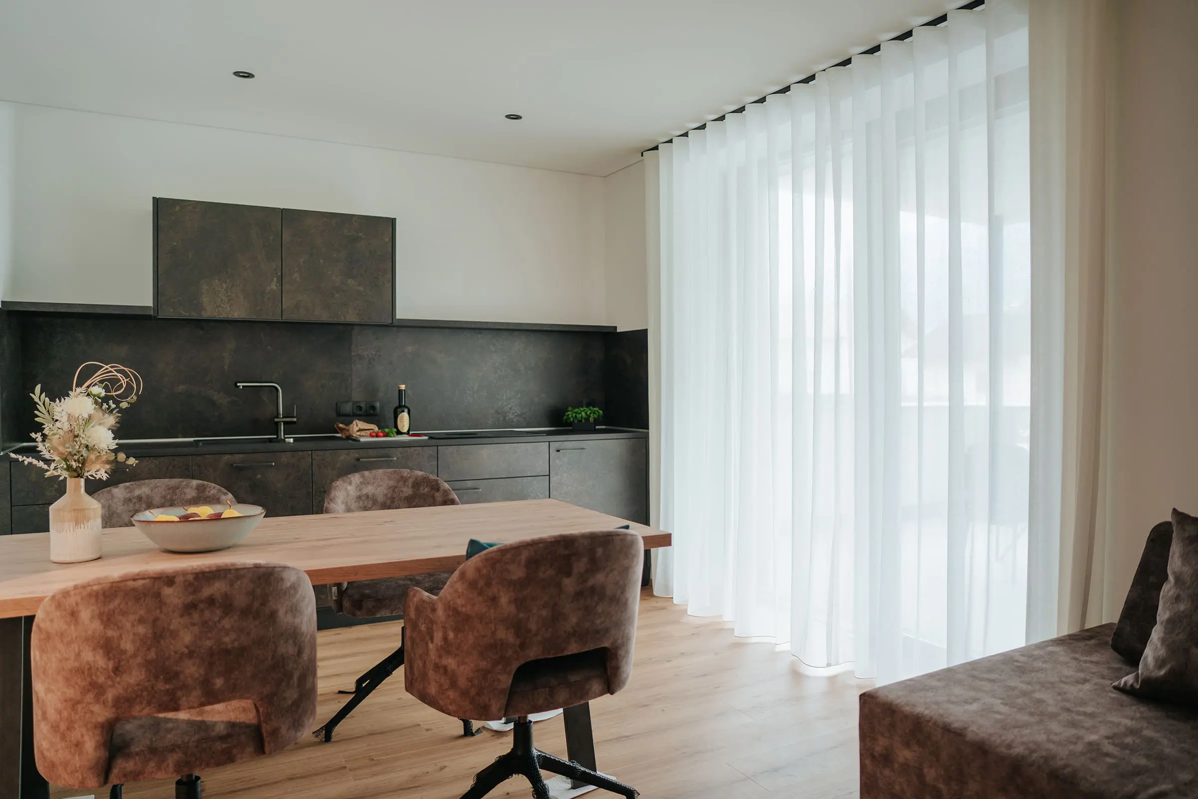 Cucina moderna e zona pranzo con tavolo di legno, sedie imbottite e grandi finestre nell'Appartamento Exclusiva dei Montoni Chalets a Prato allo Stelvio.